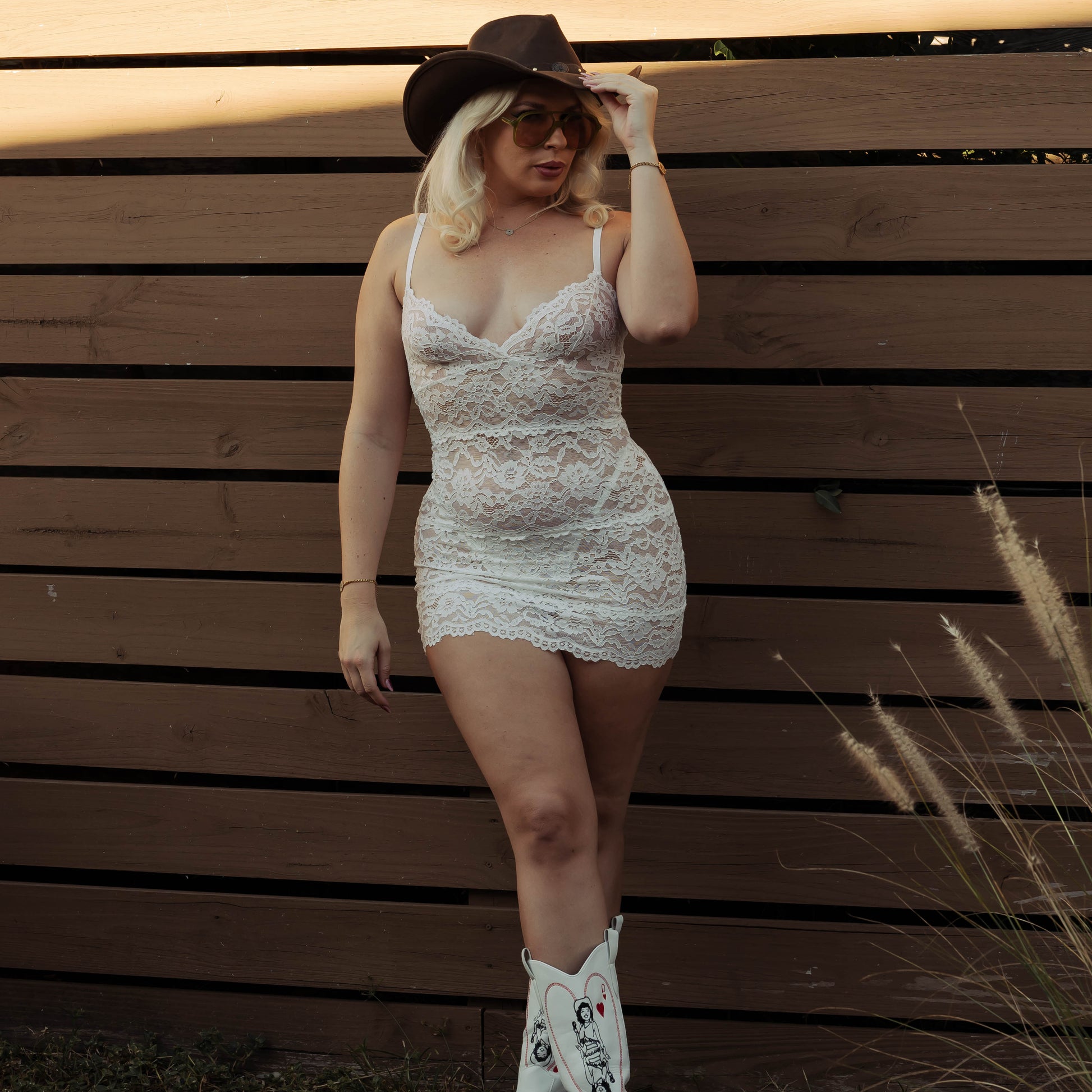 Woman in a white lace dress and black hat standing against a wooden fence.
