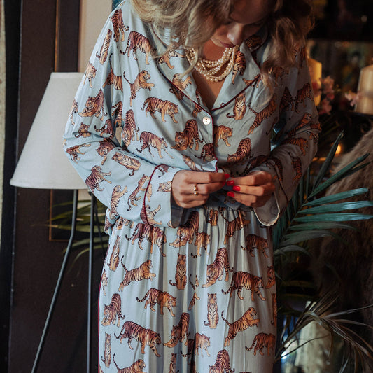 A woman wearing a light blue pajama set with an all-over orange and black tiger print, standing in a dimly lit indoor setting with plants and a lamp in the background.