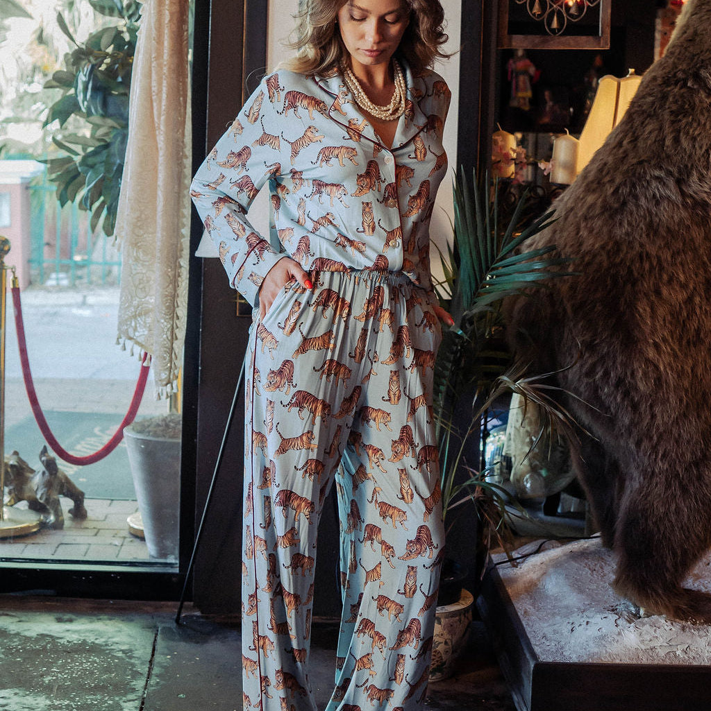A woman stands in a rustic interior wearing a light blue pajama set with an allover brown animal print. The room features a large taxidermy bear head, a patterned rug, and warm ambient lighting.