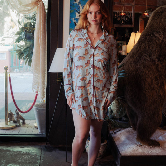 A woman stands barefoot in a rustic indoor setting wearing a long-sleeve button-up shirt dress with an allover fox print. The dress has a cream base with orange-brown and beige foxes.