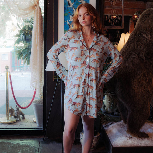 A woman stands barefoot indoors wearing a long-sleeve pajama romper with an allover orange animal print on a light background. The room has a window with sheer curtains, framed artwork, a lamp, and a large brown bear statue on a pedestal.