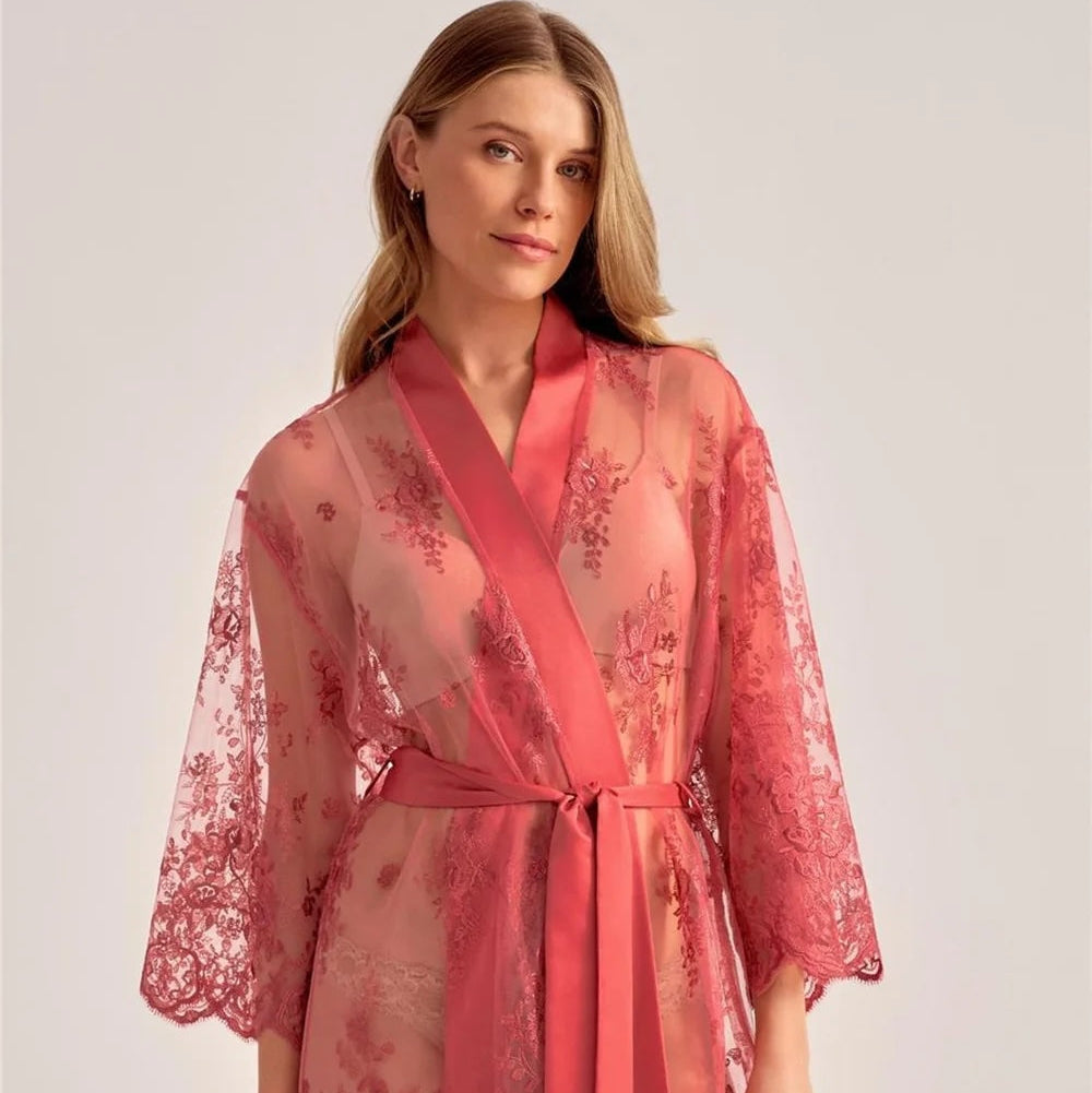 A woman wearing a sheer coral pink lace robe with a matching satin tie belt, photographed against a plain light gray studio background.