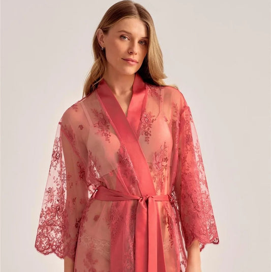 A woman wearing a sheer coral pink lace robe with a matching satin tie belt, photographed against a plain light gray studio background.