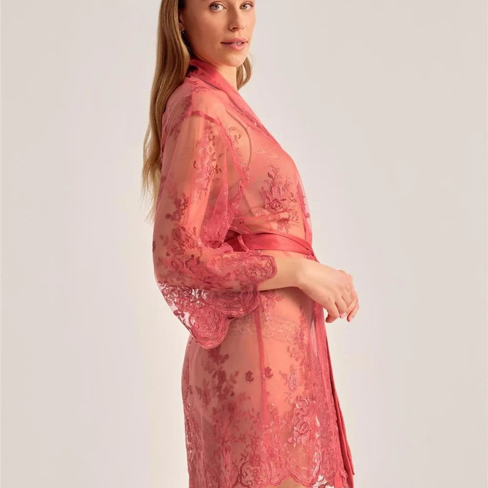A woman modeling a sheer pink lace robe with floral embroidery and a matching waist tie, photographed against a plain light gray studio background.