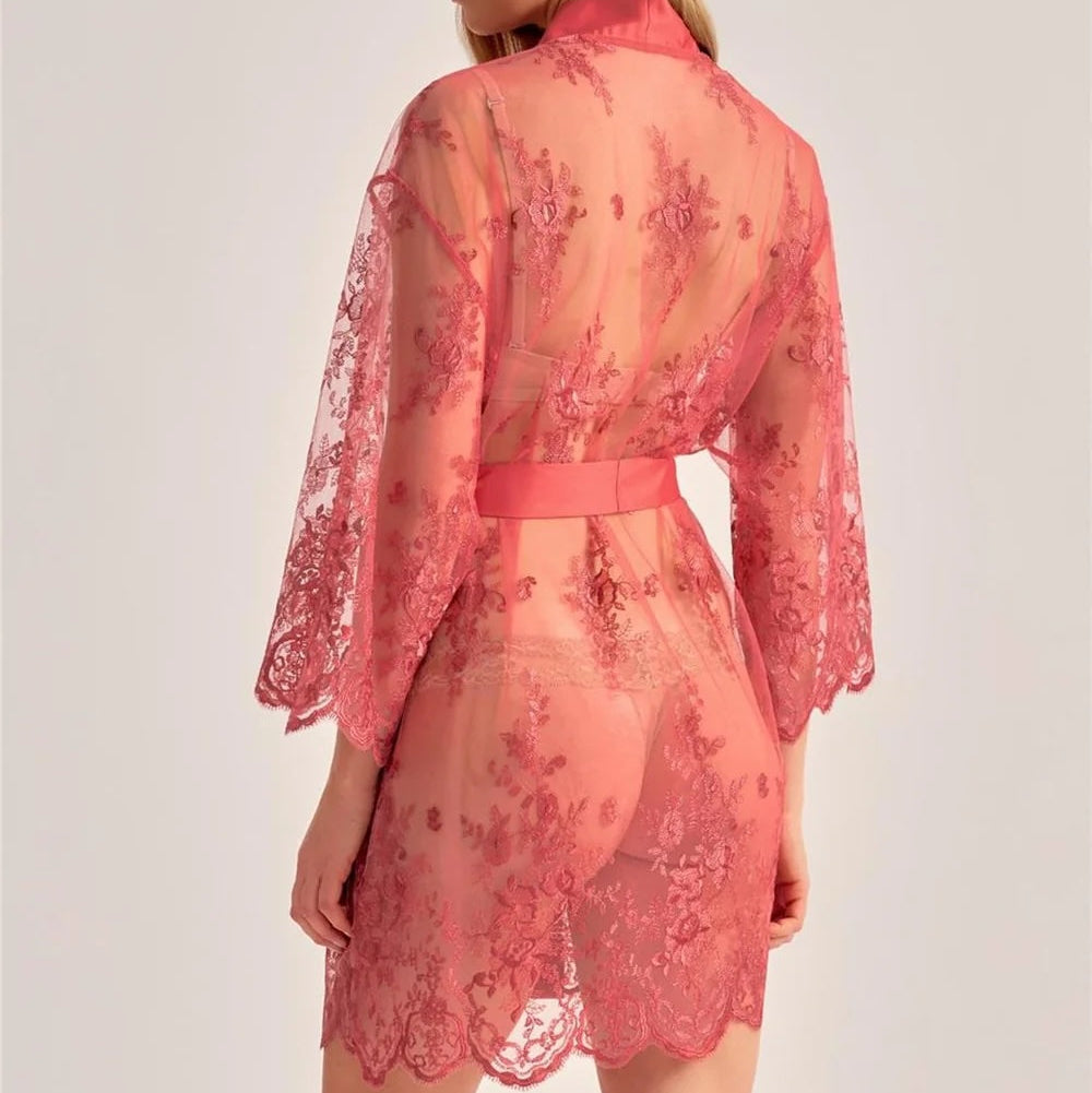 A woman shown from the back wearing a sheer coral pink lace robe with floral embroidery and a matching waist tie, against a plain light gray studio background.