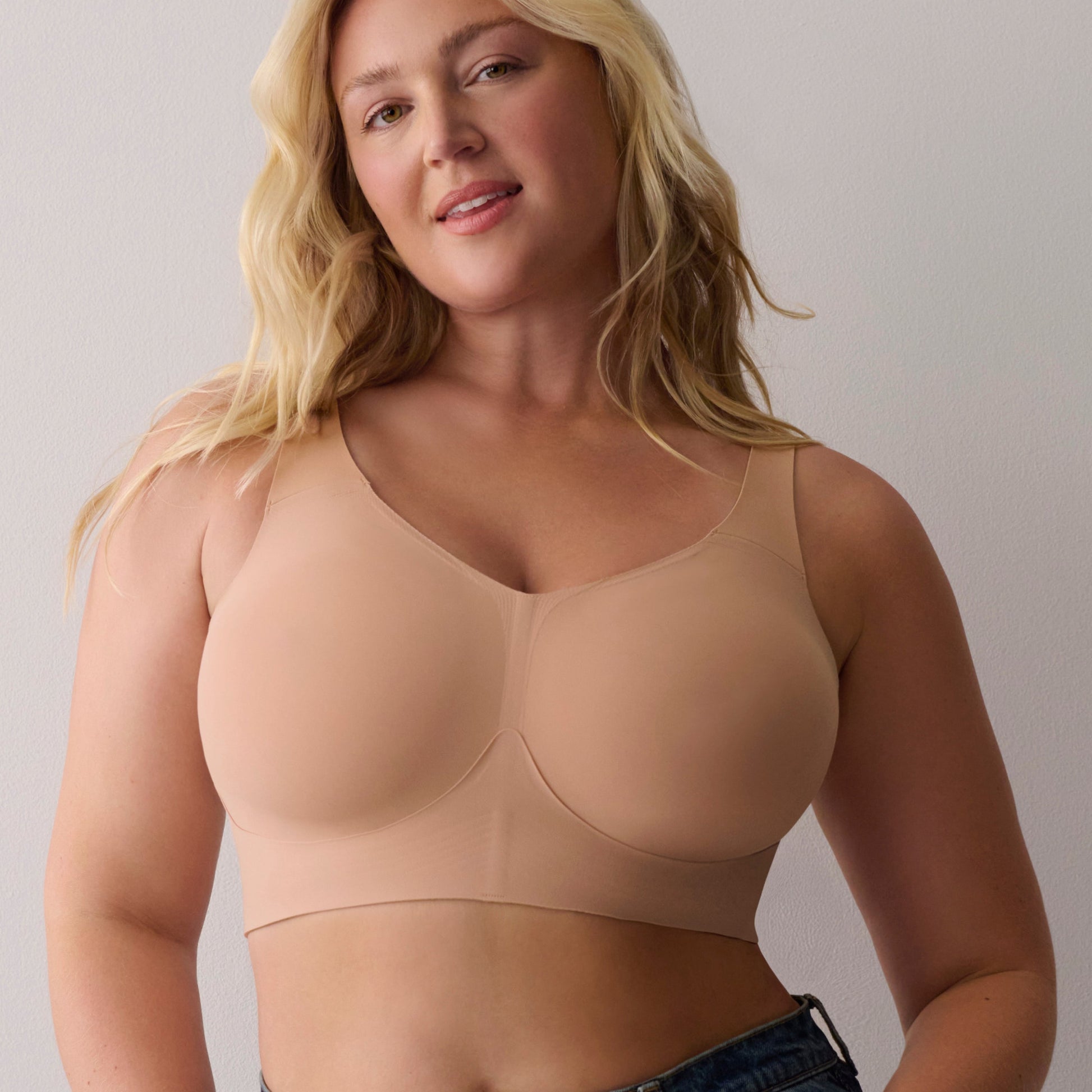 A blonde woman modeling a seamless, scoop-neck beige bralette with underwire cups, paired with high-waisted blue jeans against a neutral light gray background.