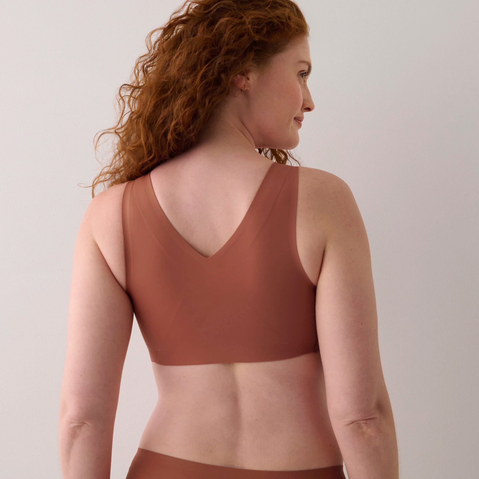 A woman with curly red hair is shown from the back wearing a solid terracotta brown bralette and matching underwear against a plain light gray background.