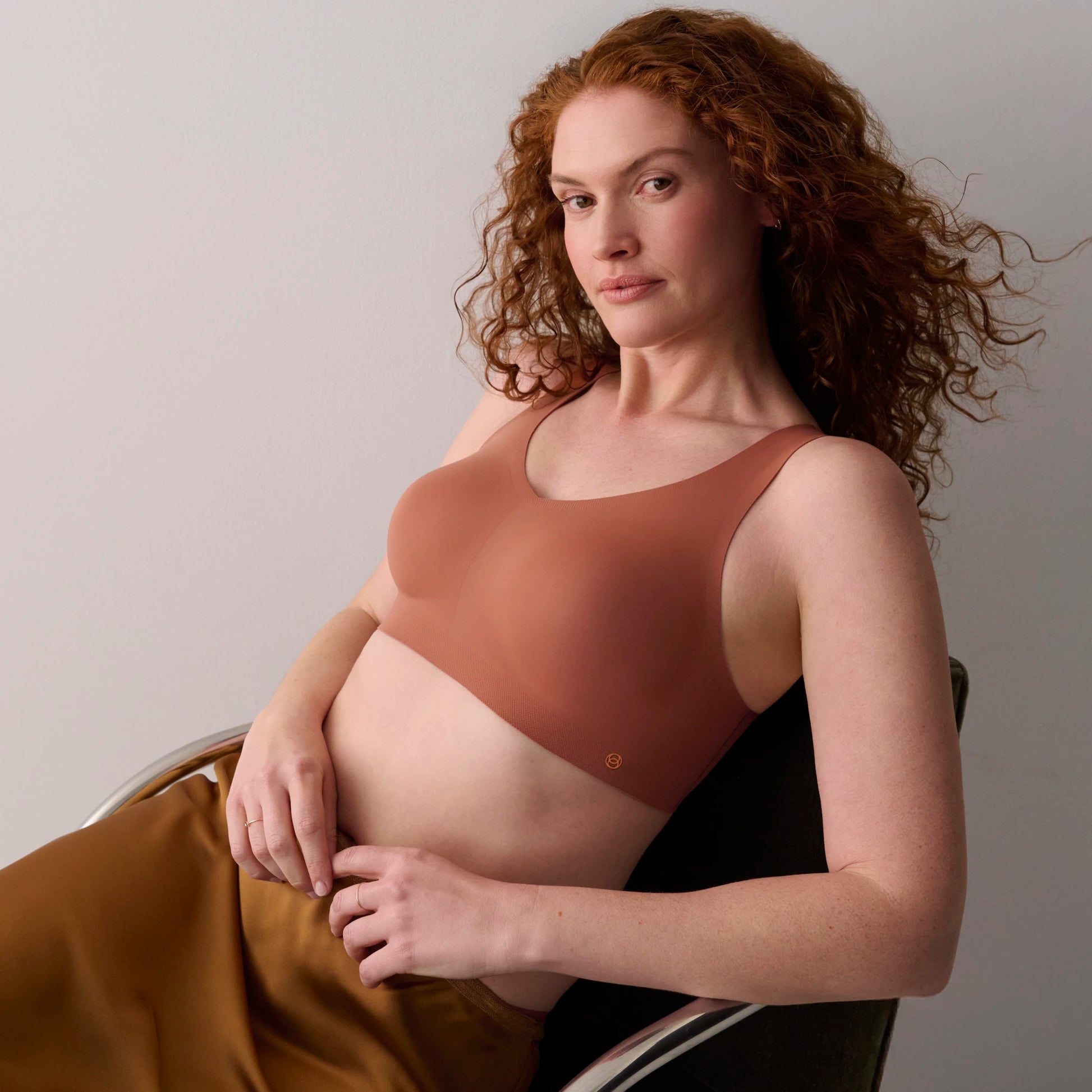 A pregnant woman with curly red hair wearing a seamless, molded maternity bra in a muted terracotta brown color, sitting in a dark chair against a neutral light gray background.