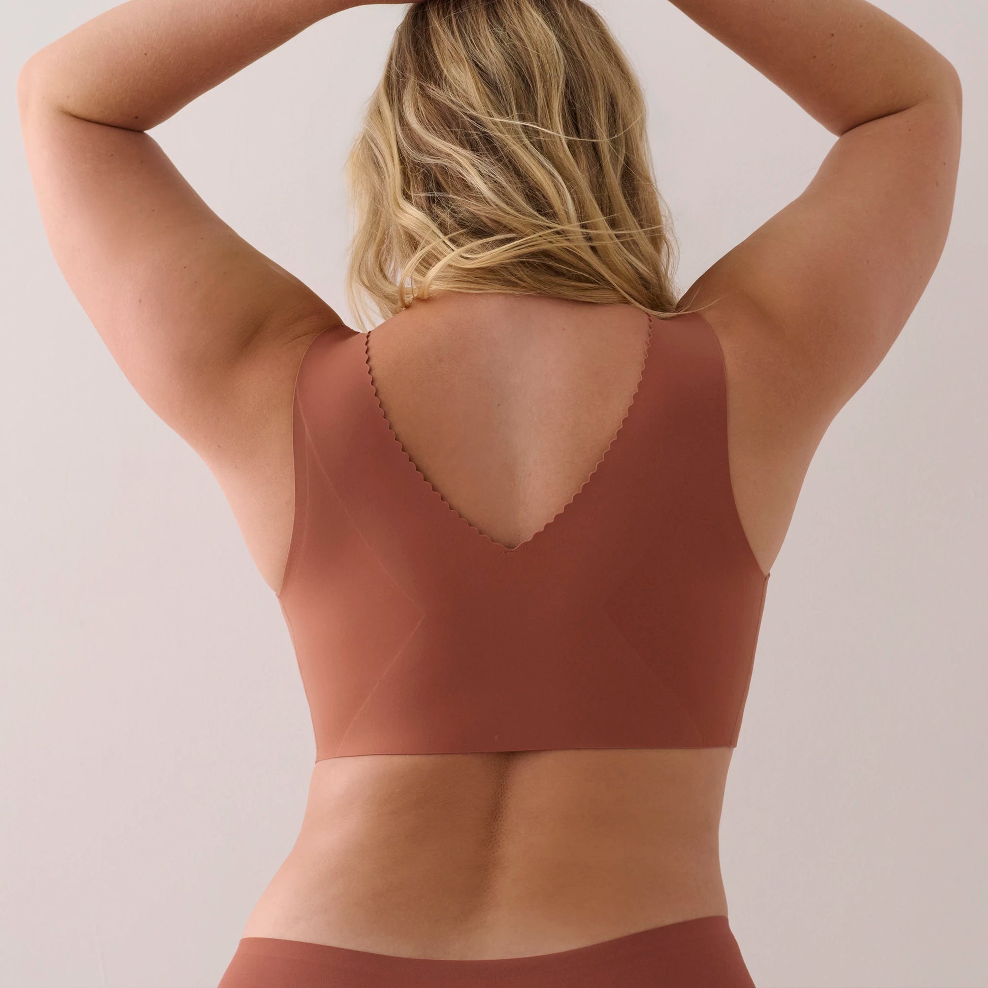 A woman viewed from behind wearing a solid terracotta brown sports bra and matching bottoms against a plain light beige studio background.