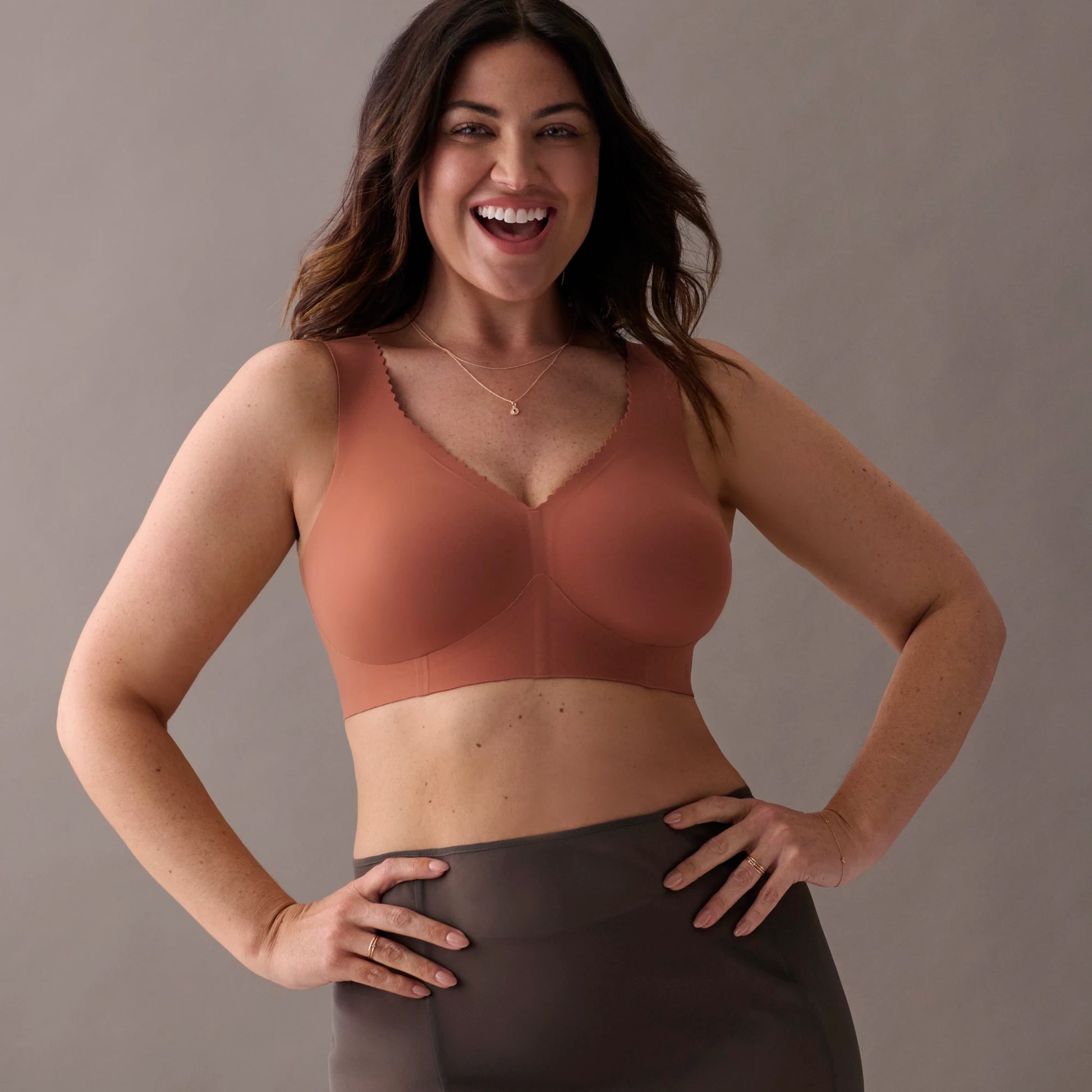 A smiling woman modeling a seamless, scoop-neck bra in a warm terracotta or rust shade, paired with dark gray high-waisted leggings, against a solid neutral gray studio background.