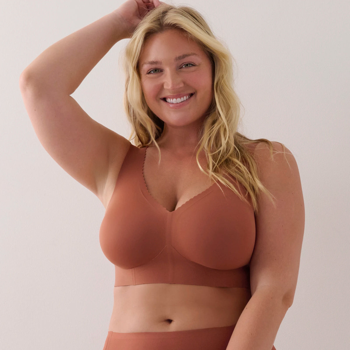 A smiling blonde woman wearing a terracotta-colored wireless bra and matching underwear against a plain light beige background.