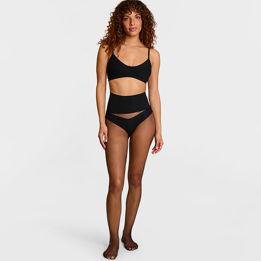 A model with curly brown hair stands against a light gray studio background wearing a black two-piece swimsuit with a bandeau-style top and high-waisted bottoms with a sheer mesh detail.