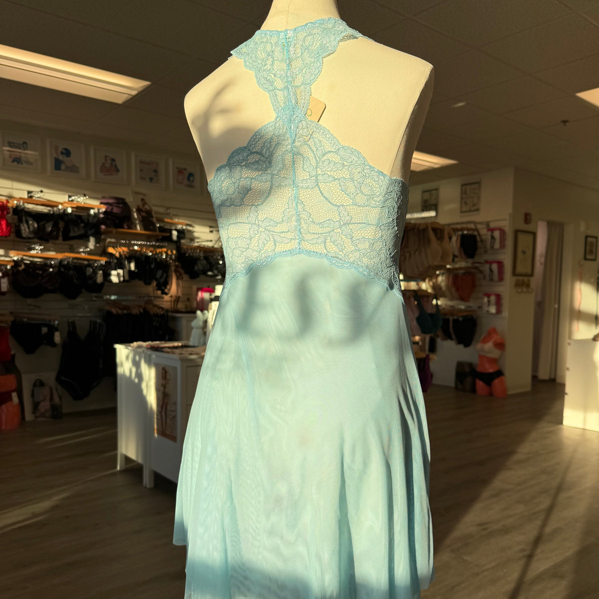 A light blue lace and satin halter dress displayed on a mannequin inside a retail store. The back view shows intricate light blue lace detailing across the upper back and neck, with a flowing satin skirt below.