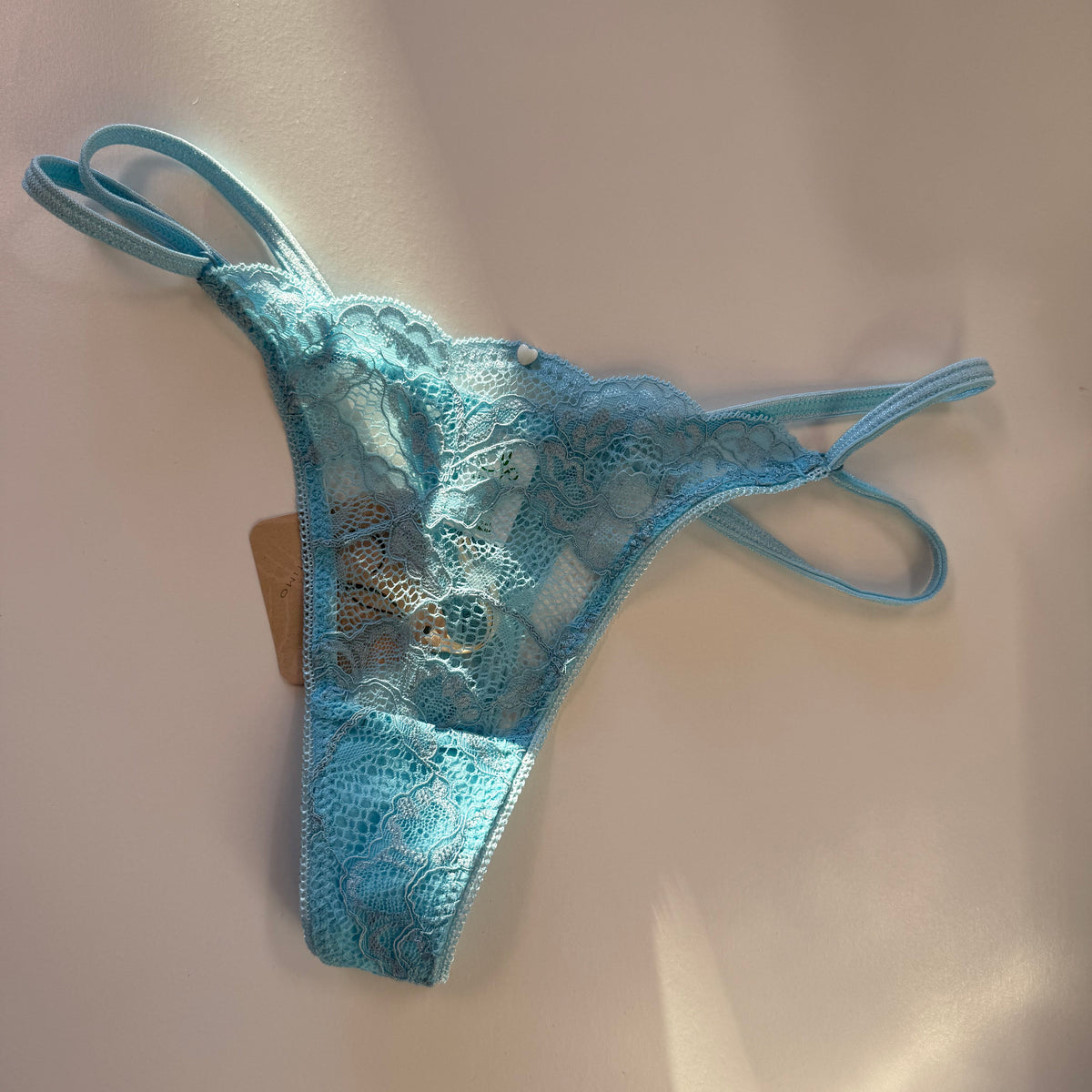 Light blue lace thong panties with thin adjustable straps, displayed on a light beige surface with a small paper tag attached.