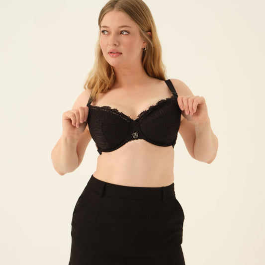 A woman modeling a black lace underwire bra and a matching black high-waisted mini skirt with side slits, set against a plain off-white studio background.