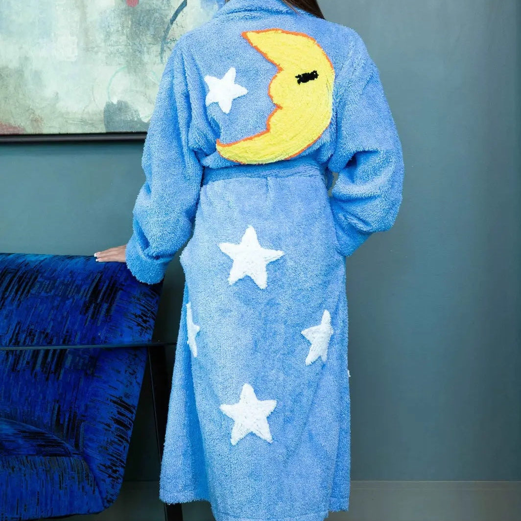 A woman viewed from behind wearing a light blue plush bathrobe with white stars and a yellow crescent moon applique on the back, standing indoors against a blue-gray wall.