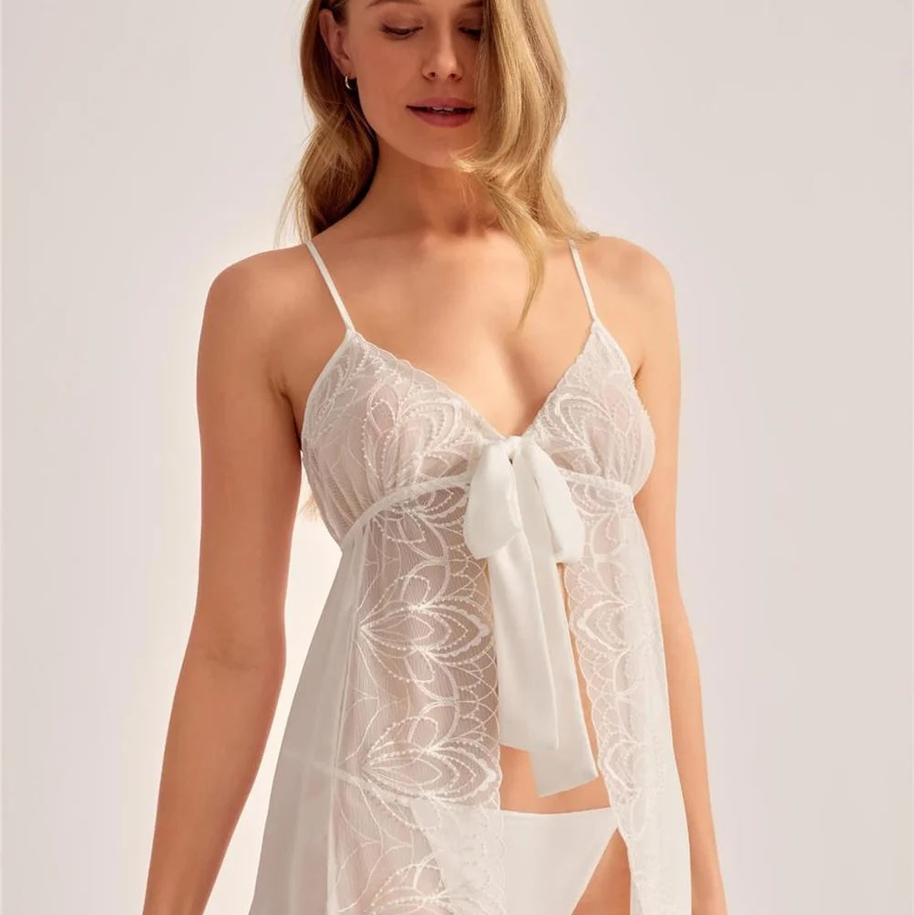 A woman modeling a white sheer lace babydoll lingerie chemise with floral embroidery and a front bow, paired with matching white underwear, against a light gray studio background.