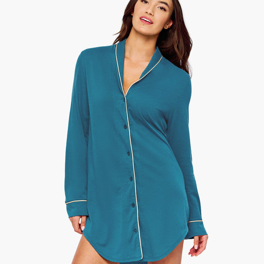 A woman modeling a teal long-sleeve button-front shirt dress with thin cream piping details on the collar, front placket, and cuffs, set against a plain white studio background.