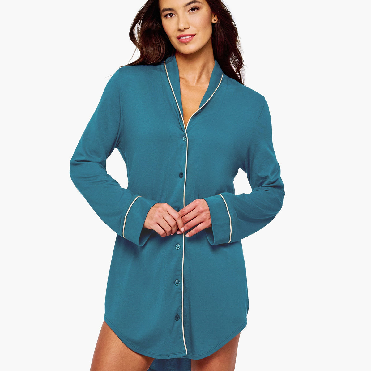 A woman modeling a teal blue long-sleeve button-front nightshirt with white piping trim on the collar, cuffs, and front placket, against a plain white studio background.