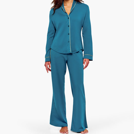 A woman modeling a two-piece teal pajama set with a long-sleeve button-front top and matching flared pants, photographed against a clean light gray studio background.