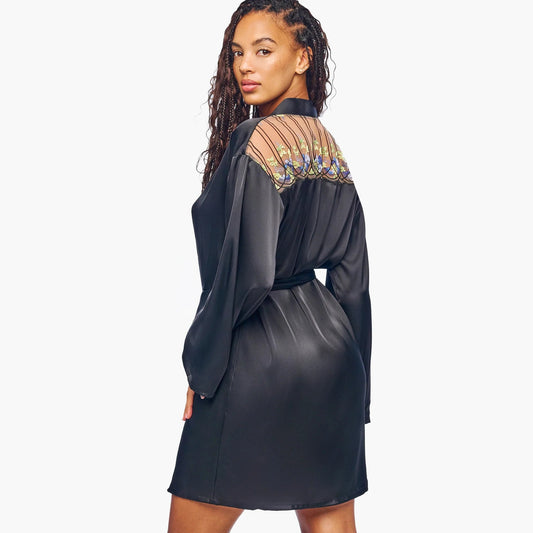 A woman modeling a black long-sleeve mini dress with sheer patterned mesh shoulder panels, shown from a rear three-quarter angle against a light gray studio background.