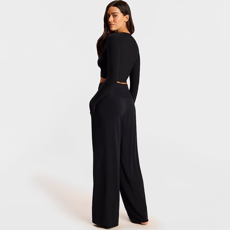 A woman modeling a black long-sleeve jumpsuit with a low back cutout, shown from a rear three-quarter angle against a light gray studio background.