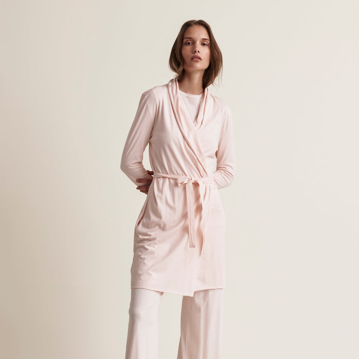 A woman modeling a light pink long-sleeve wrap robe with a tie belt and matching wide-leg pants against a plain beige studio background.