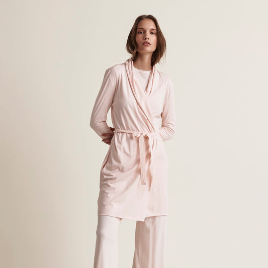 A woman modeling a light pink long-sleeve wrap robe with a tie belt and matching wide-leg pants against a plain beige studio background.
