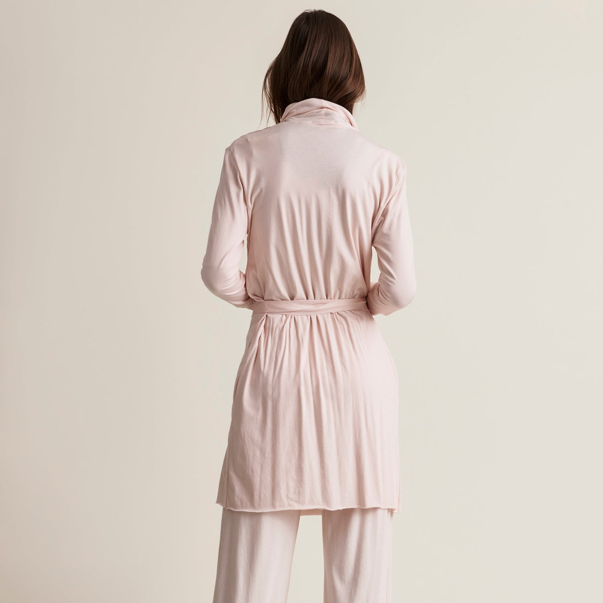 A model shown from the back wearing a light blush pink long-sleeve belted shirt and matching wide-leg pants against a solid warm beige studio background.