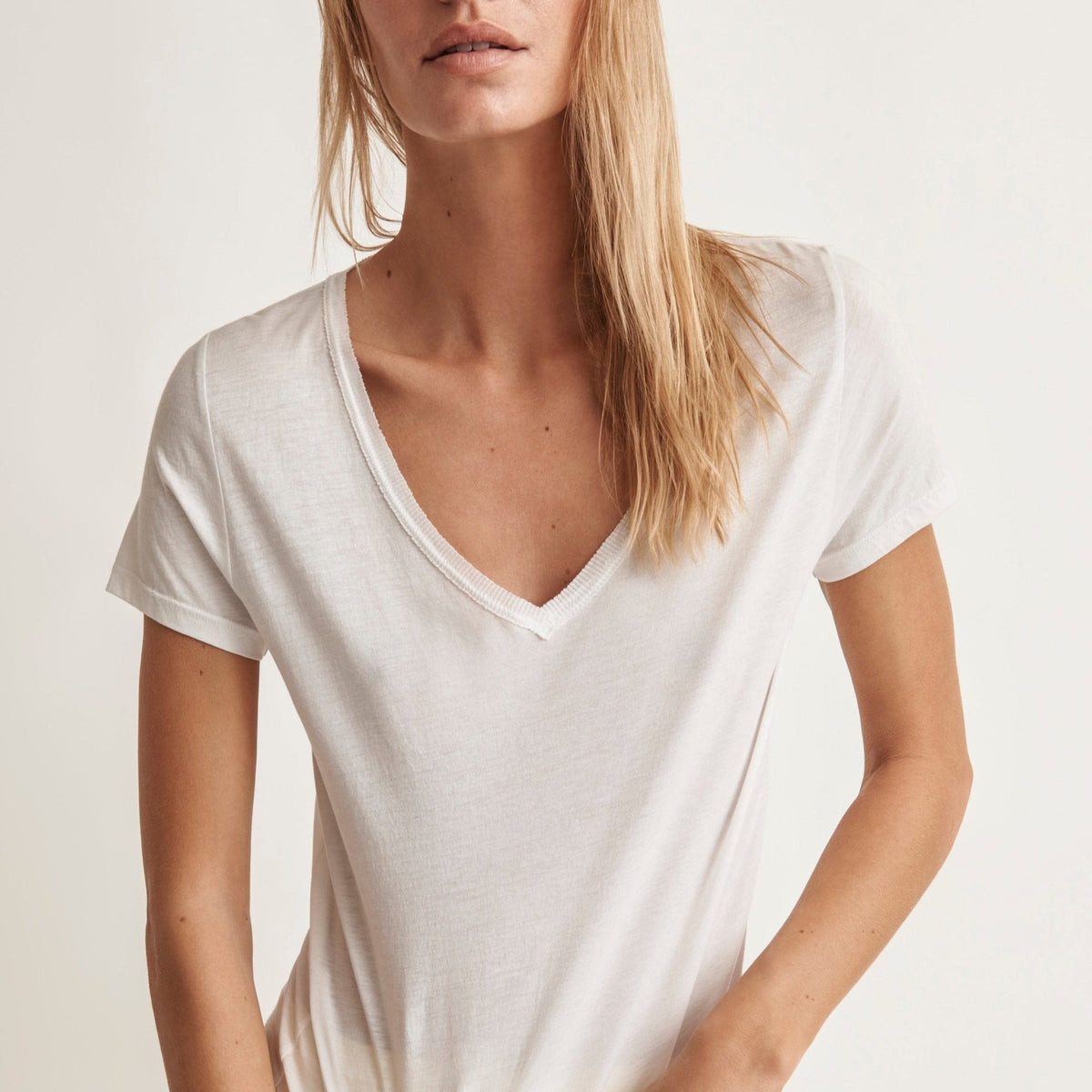 A woman wearing a short-sleeve V-neck t-shirt in a warm off-white or cream color, paired with light yellow pants, against a plain light background.
