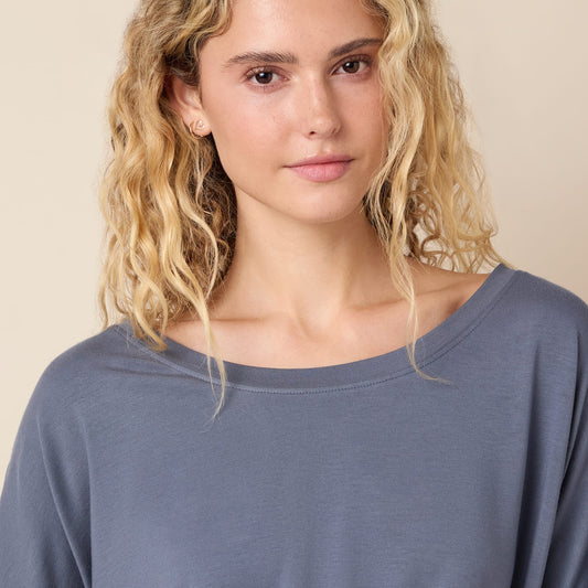 A woman with blonde curly hair wearing a slate blue long-sleeve top, photographed against a solid beige background.