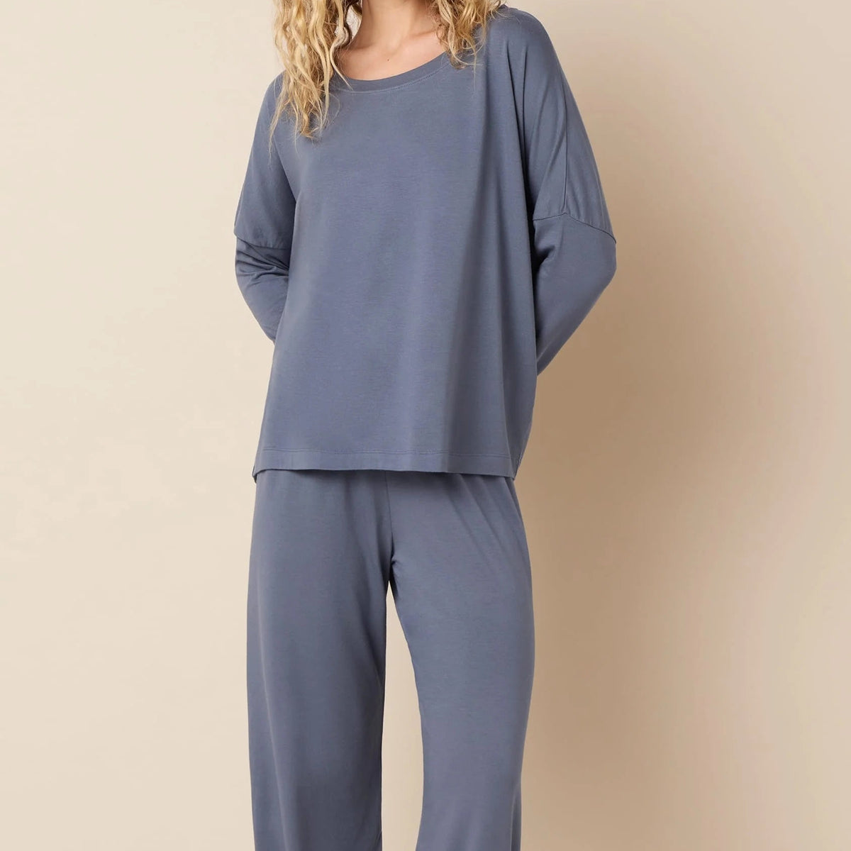 A woman modeling a two-piece loungewear set consisting of a loose long-sleeve top and matching wide-leg pants in a dusty slate blue color, set against a solid beige studio background.