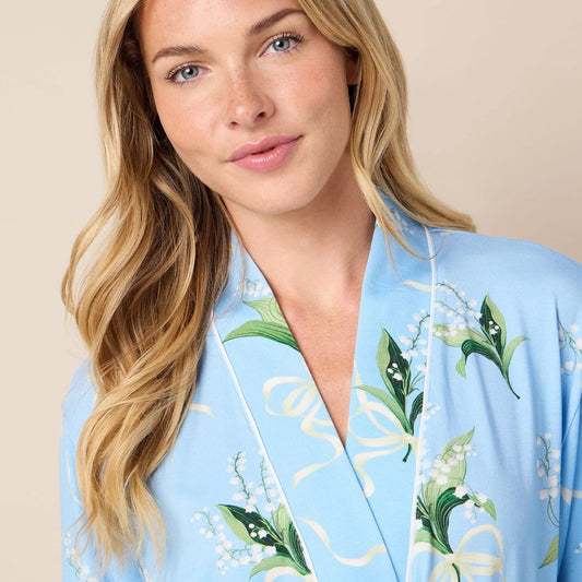 A woman wearing a light blue kimono-style robe with a green and white floral vine print, posed against a solid beige background.