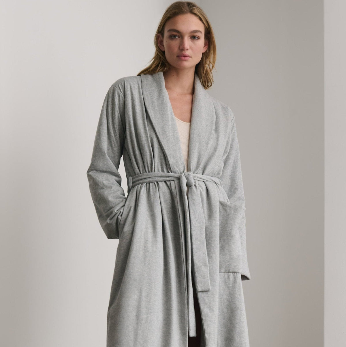 A woman wearing a light gray heathered long robe with a tie belt, standing in a minimalist room with a light gray wall.