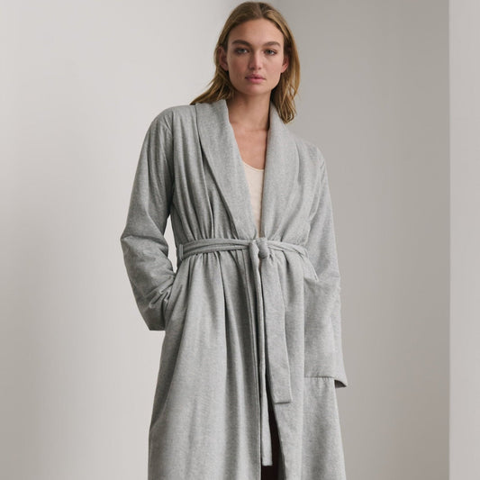 A woman wearing a light gray heathered long robe with a tie belt, standing in a minimalist room with a light gray wall.